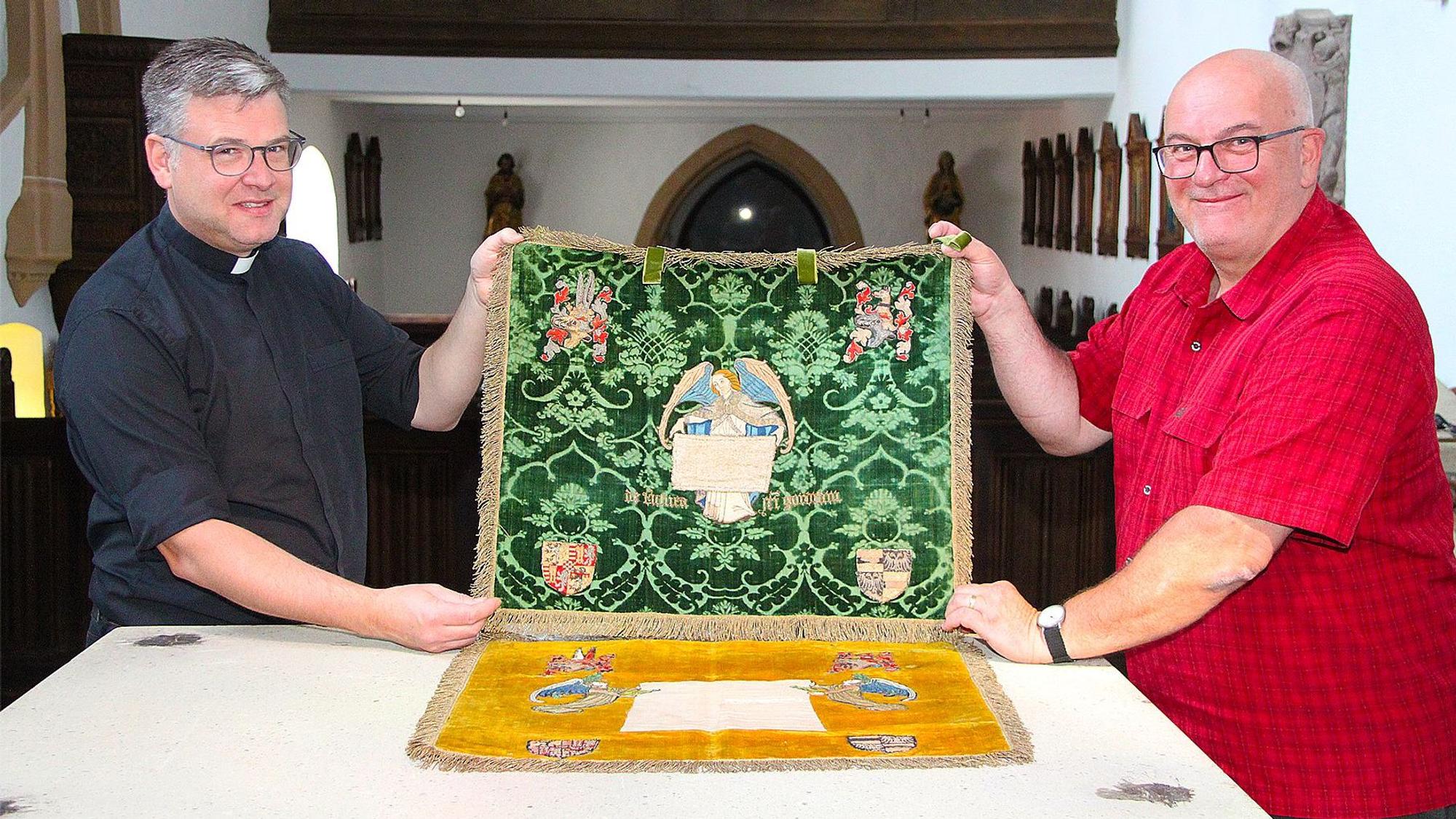 Zwei Männer, Pfarrer Andres Züll (links im schwarzen Hemd) und Markus Schmitz (rechts im roten Hemd) halten in der Pfarrkirche von Blankenheim zwei Tuchreliquien fest, die zum Kirchenschatz gehören.