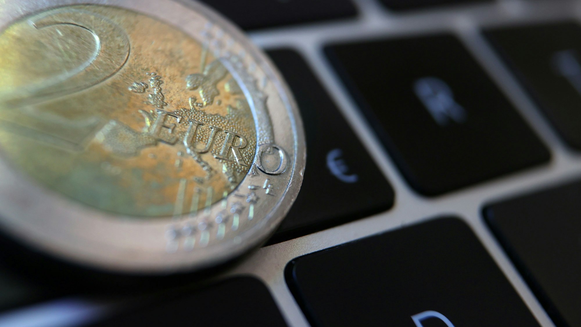 Eine Zwei-Euro-Münze liegt auf der Tastatur eines Laptops neben einem Eurozeichen (Symbolfoto).
