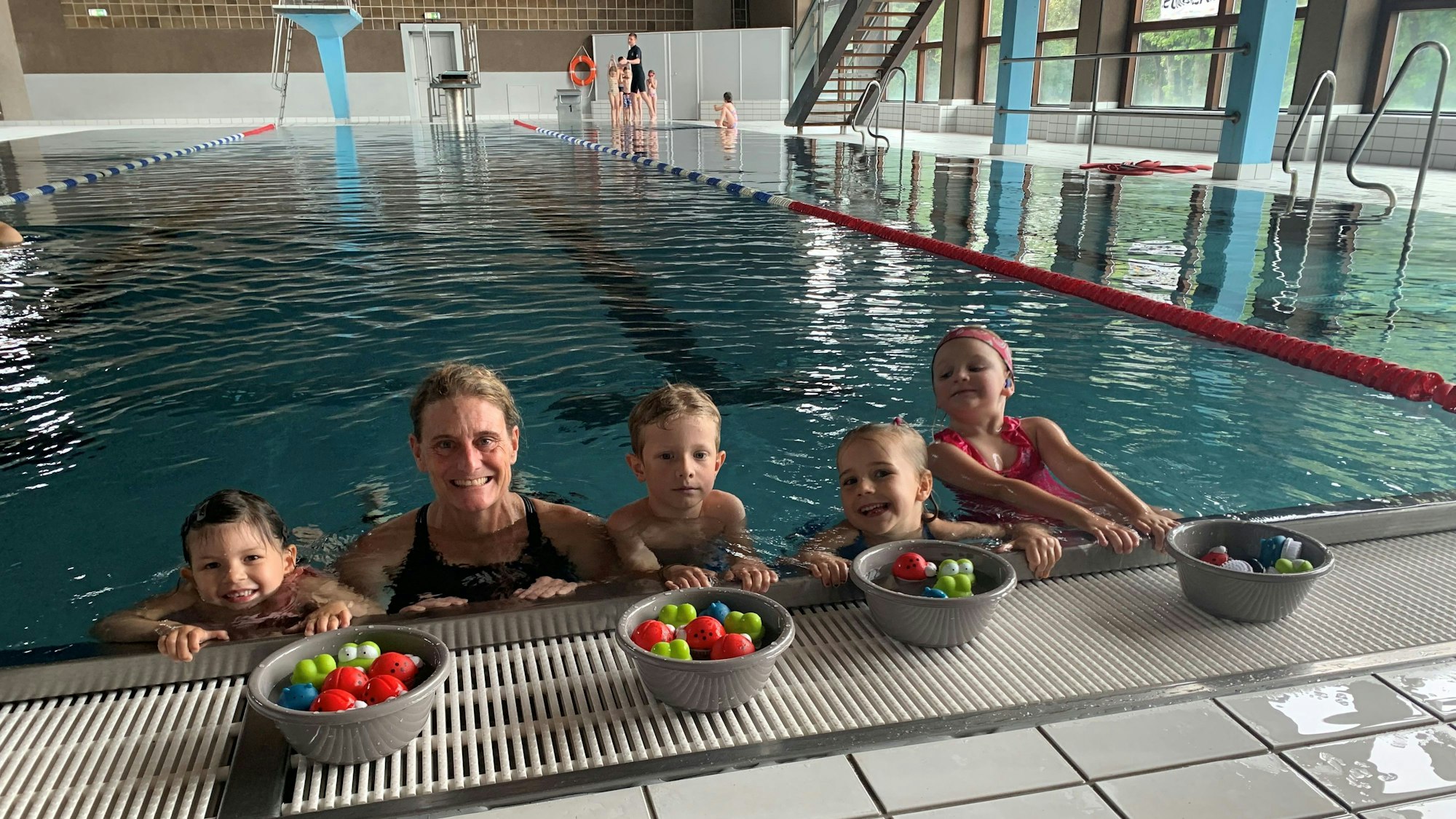 Die Schwimmlehrerin Carole Schifflers ist mit einigen Kindern im Wasser, am Beckenrand. Vor sich haben sie Schüsseln mit bunten Bällen. Im Hintergrund das Schwimmbad.
