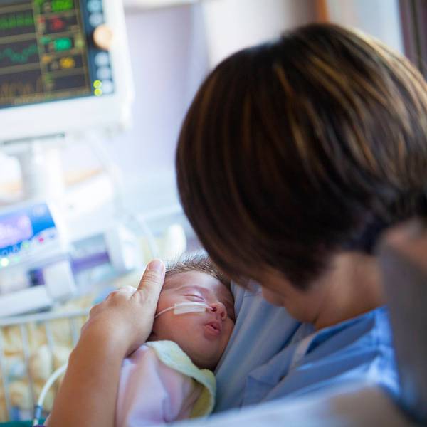 Ein Baby mit einer Sonde in der Nase liegt im Arm einer Frau. Im Hintergrund sind Monitore und medizinische Geräte zu sehen.