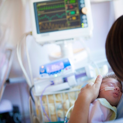 Ein Baby mit einer Sonde in der Nase liegt im Arm einer Frau. Im Hintergrund sind Monitore und medizinische Geräte zu sehen.