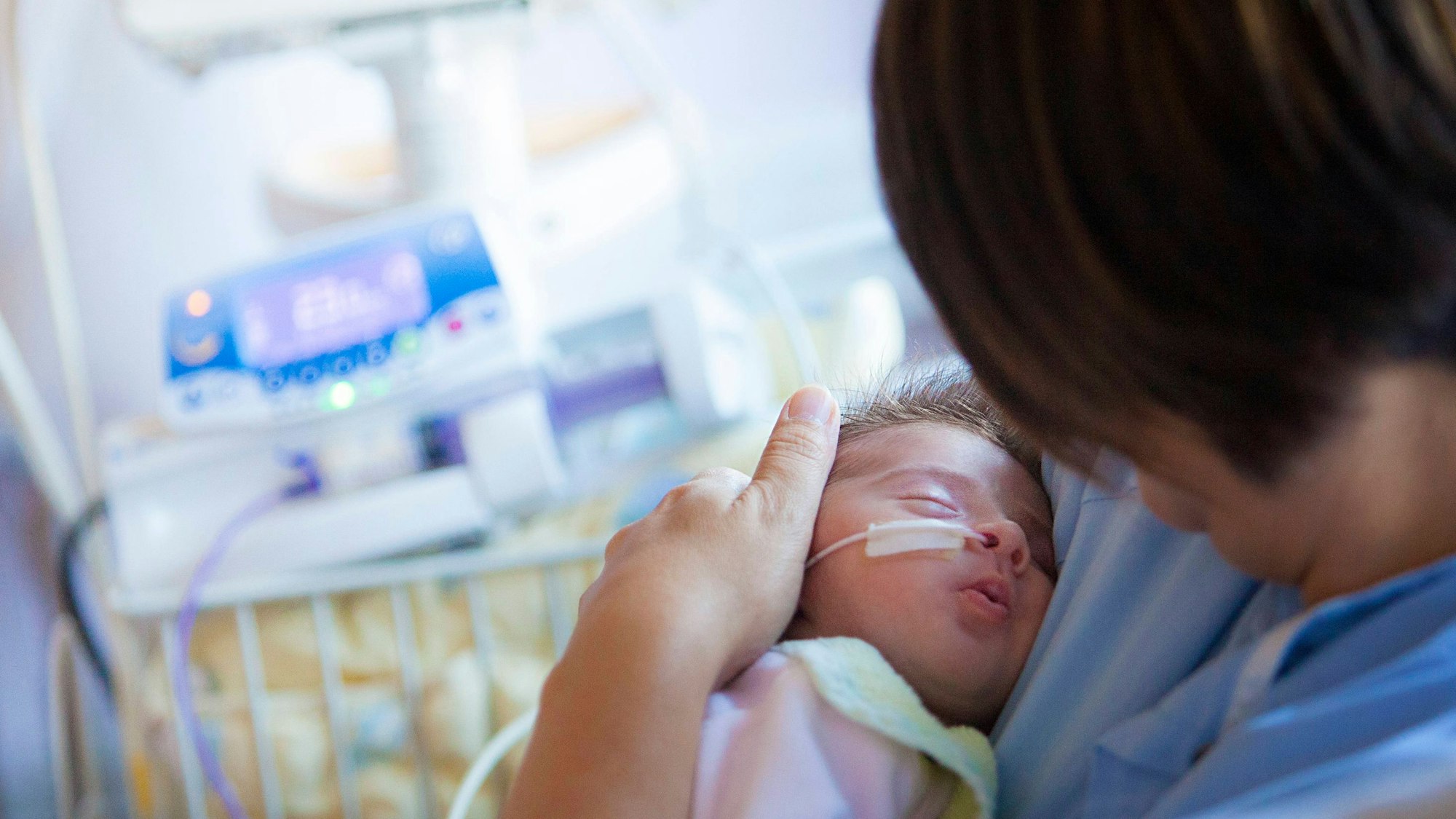 Ein Baby mit einer Sonde in der Nase liegt im Arm einer Frau. Im Hintergrund sind Monitore und medizinische Geräte zu sehen.