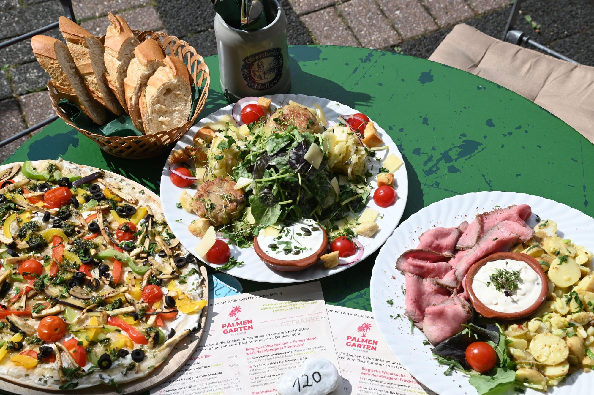 Tellergerichte sind zu sehen: ein bunt belegter Flammkuchen, rosa Roastbeefscheiben mit Kartoffelsalat und ein weiterer Teller mit Salaten.
