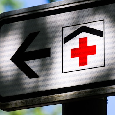 Ein Piktogramm mit einem roten Kreuz auf einem Straßenschild weist den Weg zu einem Krankenhaus.
