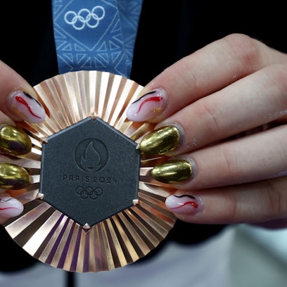Die Hände einer Frau präsentieren eine olympische Bronzemedaille an blauem Band. Die Fingernägel der Dame sind in unterschiedlichen Farben, metallisch und rosa, rot, schwarz lackiert.