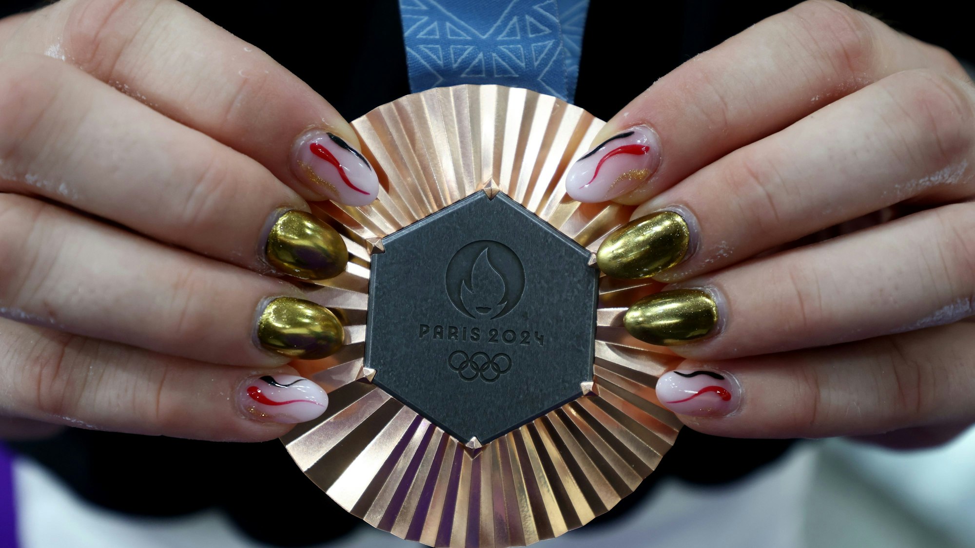 Die Hände einer Frau präsentieren eine olympische Bronzemedaille an blauem Band. Die Fingernägel der Dame sind in unterschiedlichen Farben, metallisch und rosa, rot, schwarz lackiert.