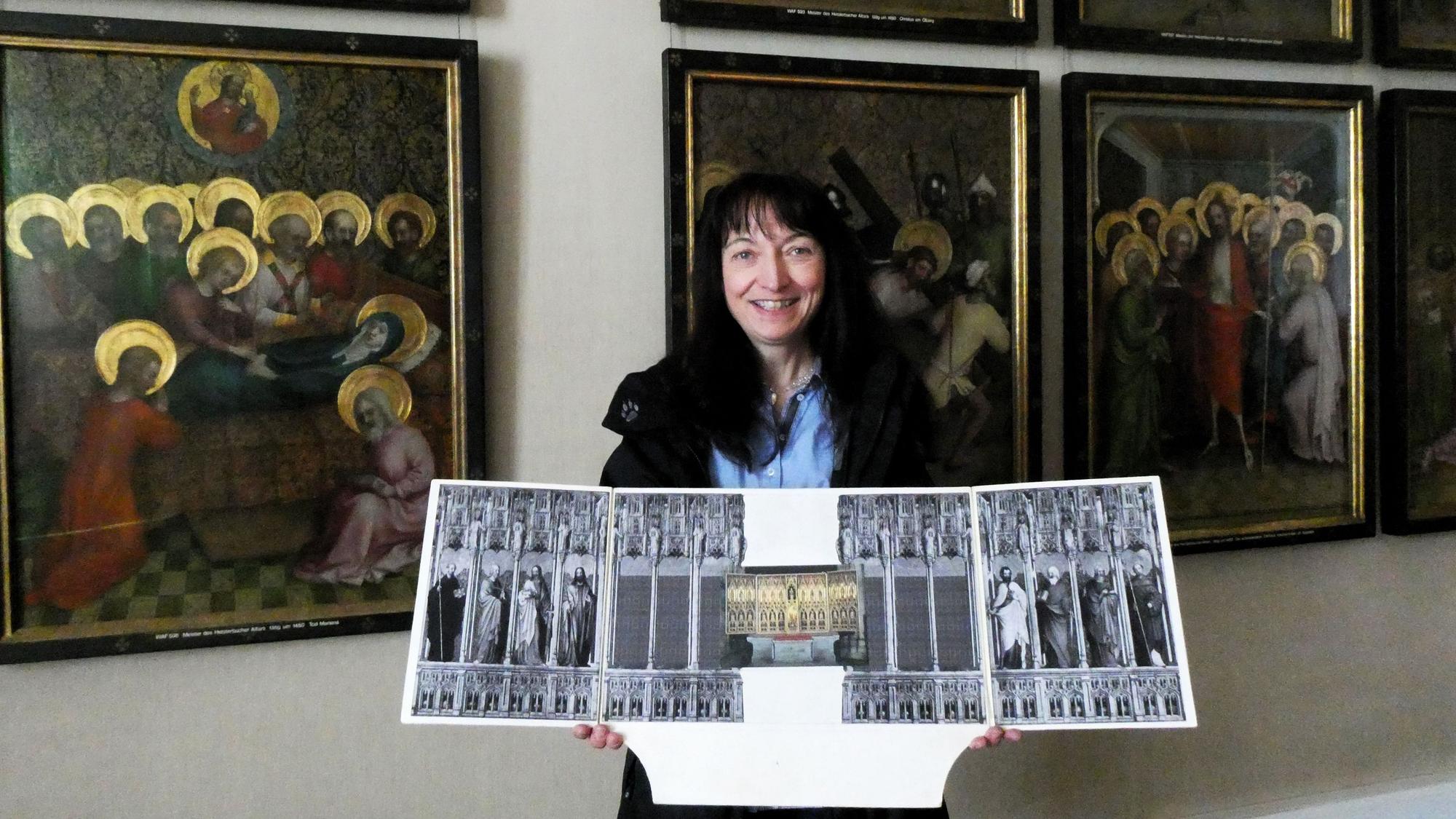 Eine Frau steht vor Gemälden und hält die Darstellung eines Altars in den Händen.