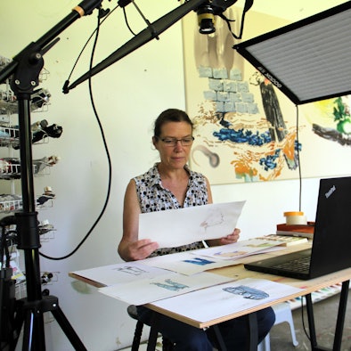 Eine Frau sitzt an einem Tisch mit vielen Zeichnungen, vor ihr ein Laptop.