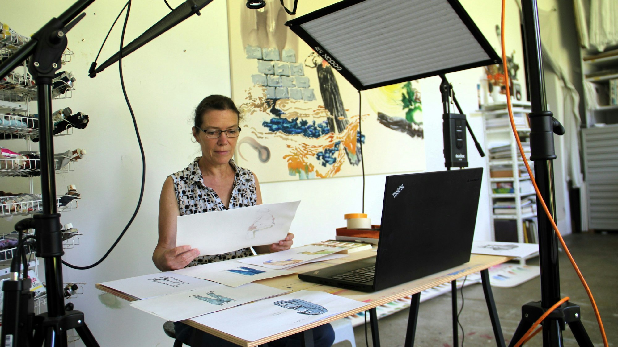 Eine Frau sitzt an einem Tisch mit vielen Zeichnungen, vor ihr ein Laptop.