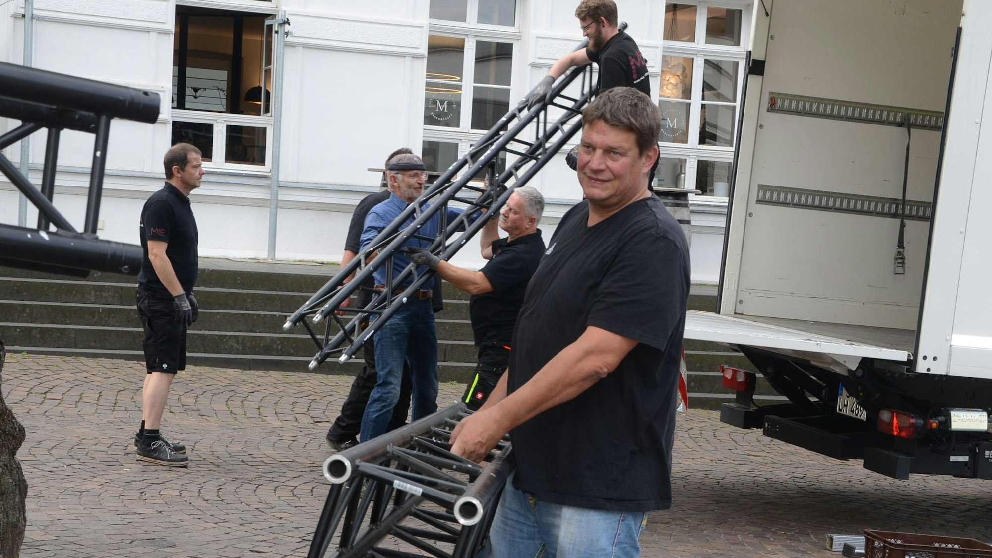 Ein Mann trägt einen Pfosten für eine Bühne.Aus einem Lastwagen werden weitere Bühnenteile herausgehoben.