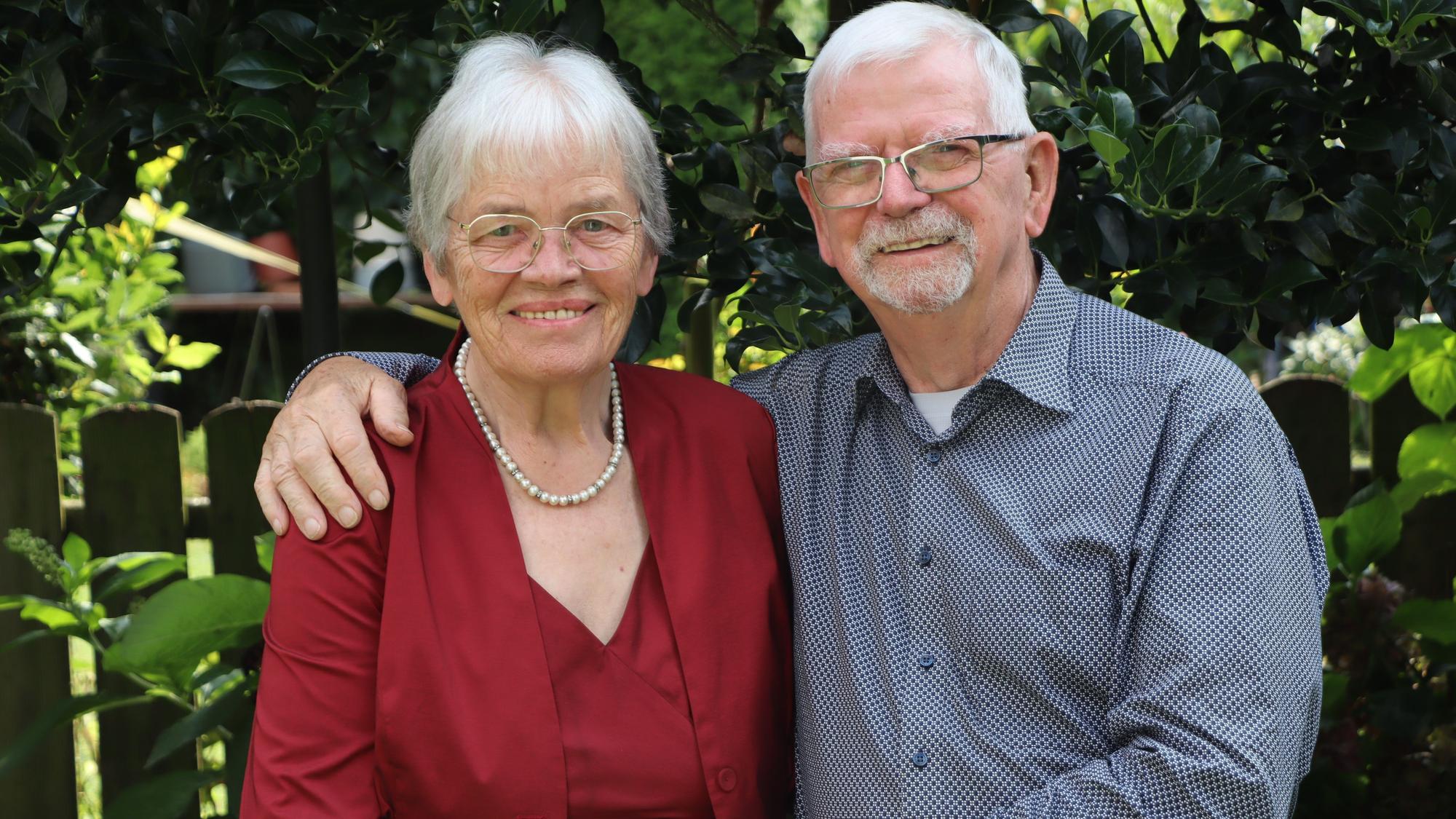 Ursula Abeln trägt eine Brille, eine rote Bluse und eine Perlenkette. Gerhard Abeln hat seinen Arm um seine Frau gelegt, er trägt eine Brille und ein blaues, gepunktetes Hemd. Im Hintergrund ein Garten.