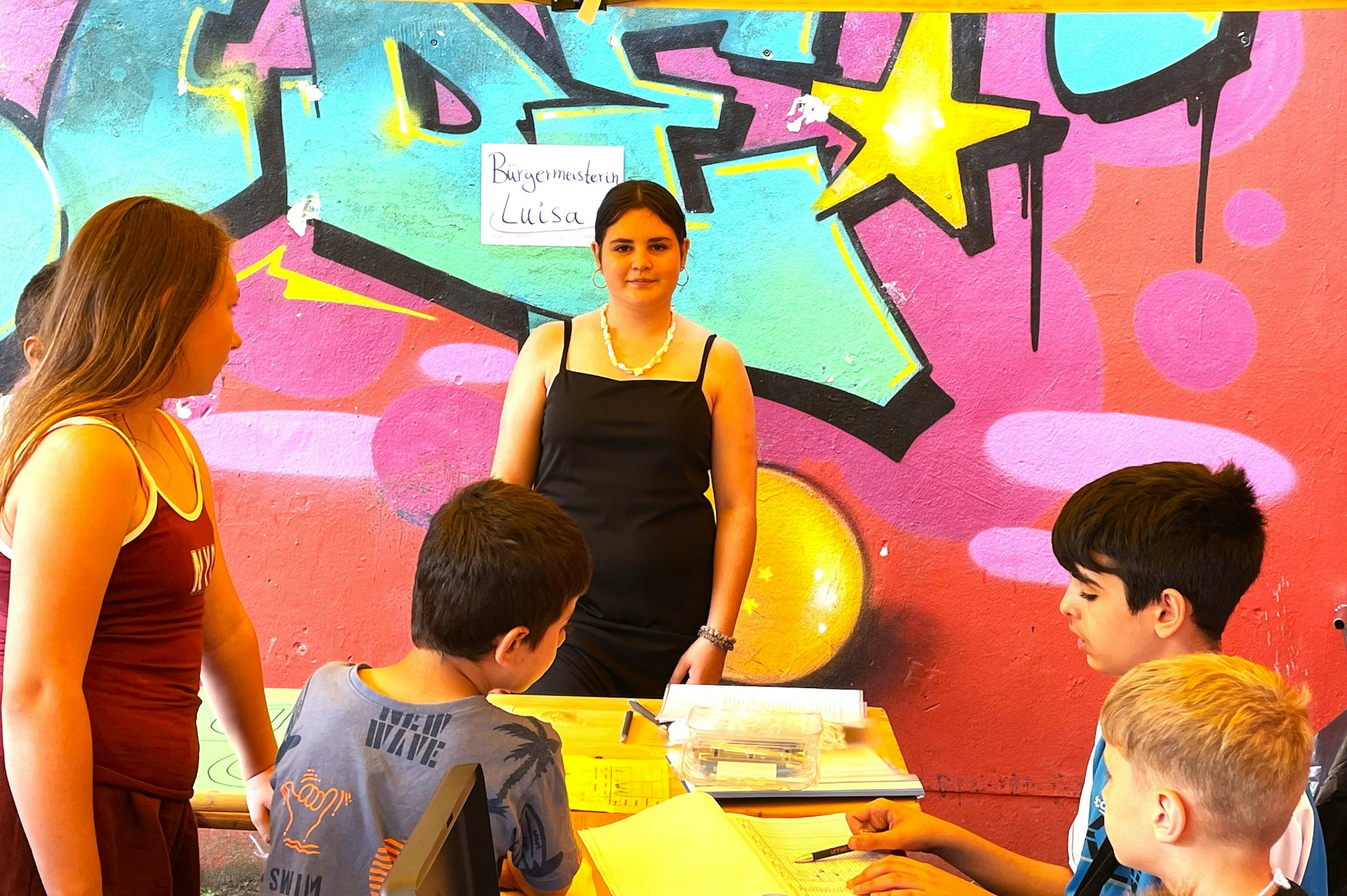 Eine 13-jährige Teilnehmerin der Kinderstadt wurde zur Bürgermeisterin gewählt, sie steht vor einer Graffiti-Wand, vor ihr sitzen Jugendliche an einem Tisch.