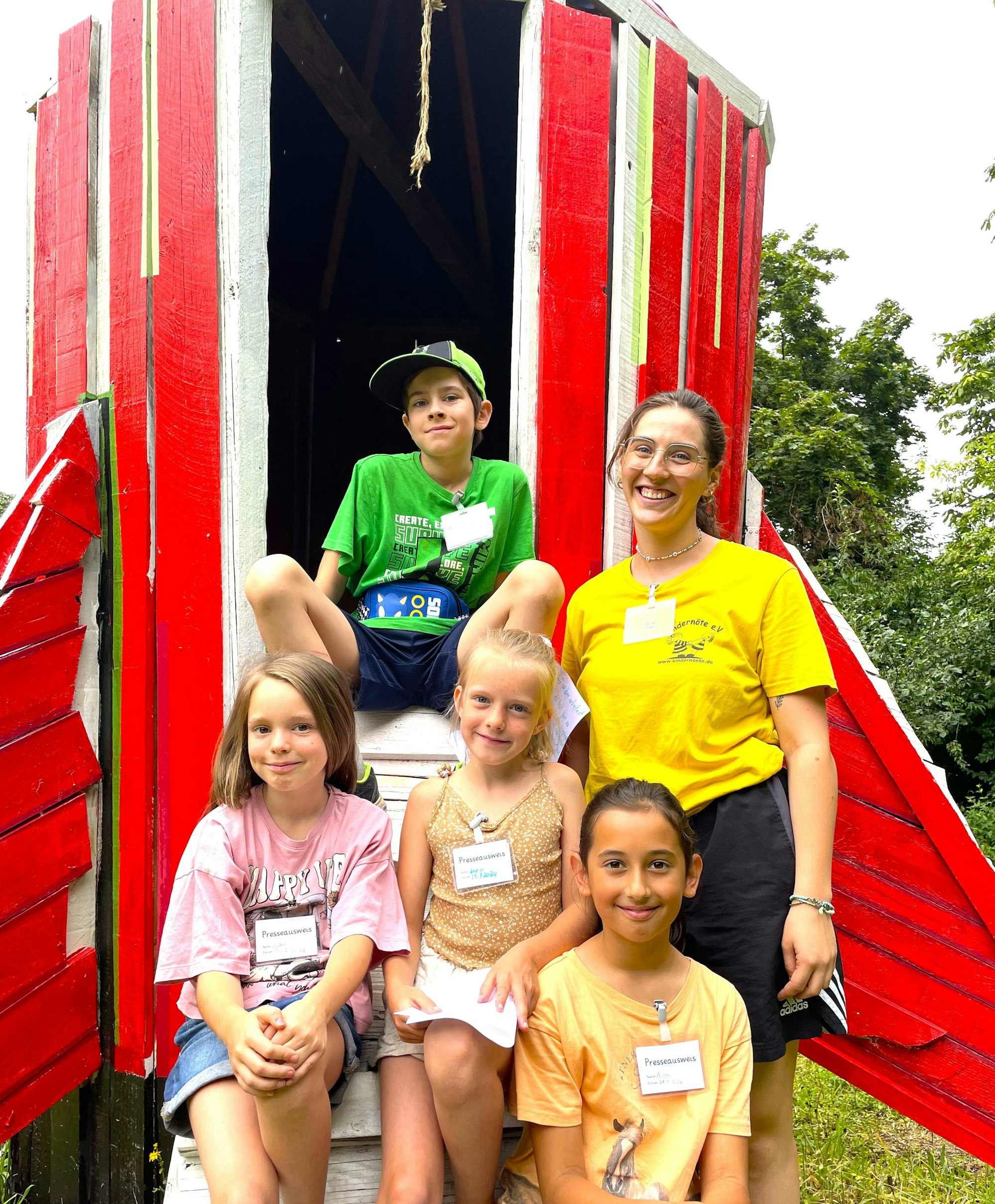 Drei Mädchen und ein Junge sitzen auf der Leiter eines Spielplatzgerätes, sie gehören zum Reportteam des Feriencamps. Neben ihnen steht eine Betreuerin die Vereins Kindernöte e.V.
