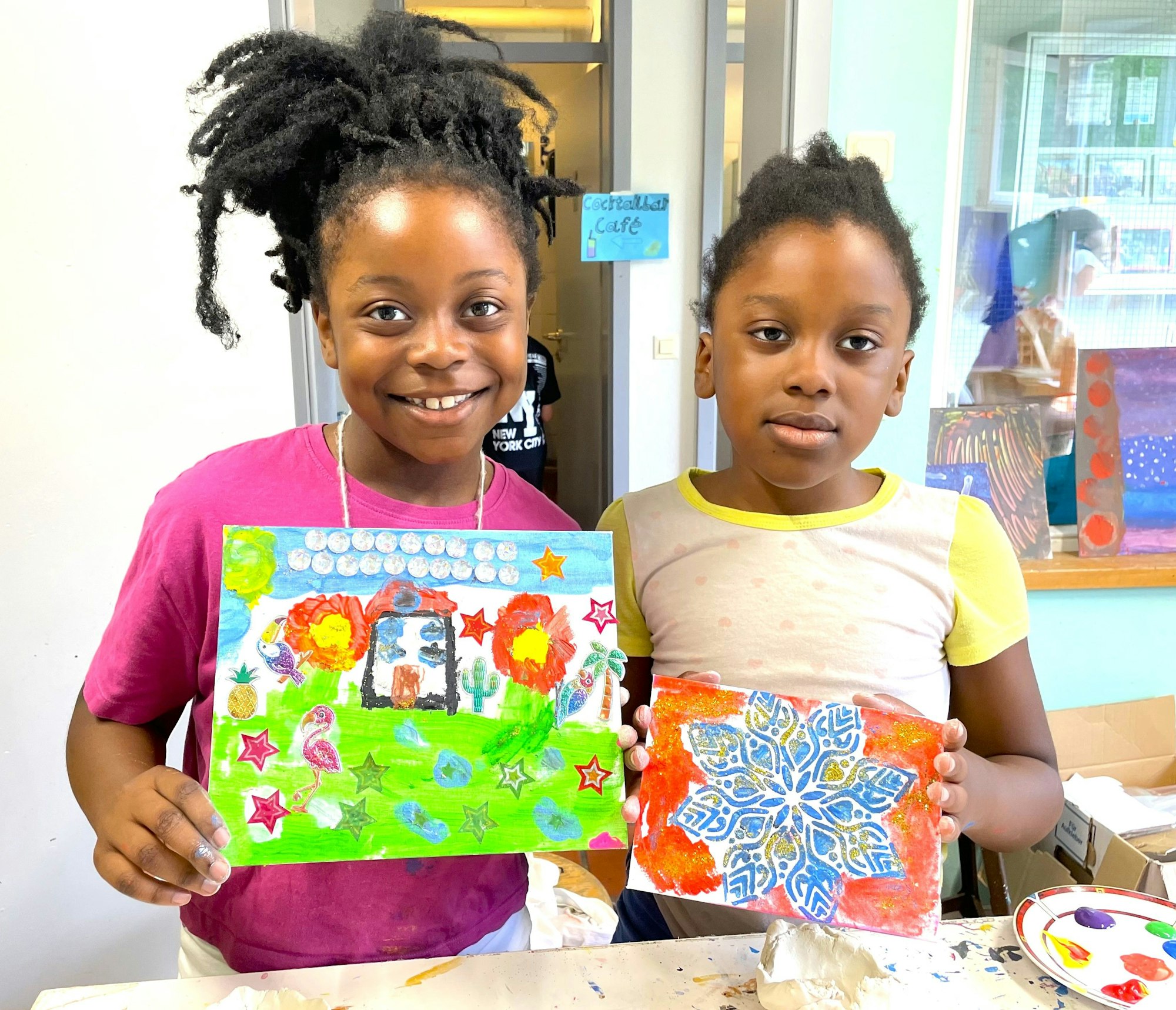 Zwei junge Mädchen halten stolz ihre bunten Gemälde in der Hand, die sie in der Malwerkstatt angefertigt haben