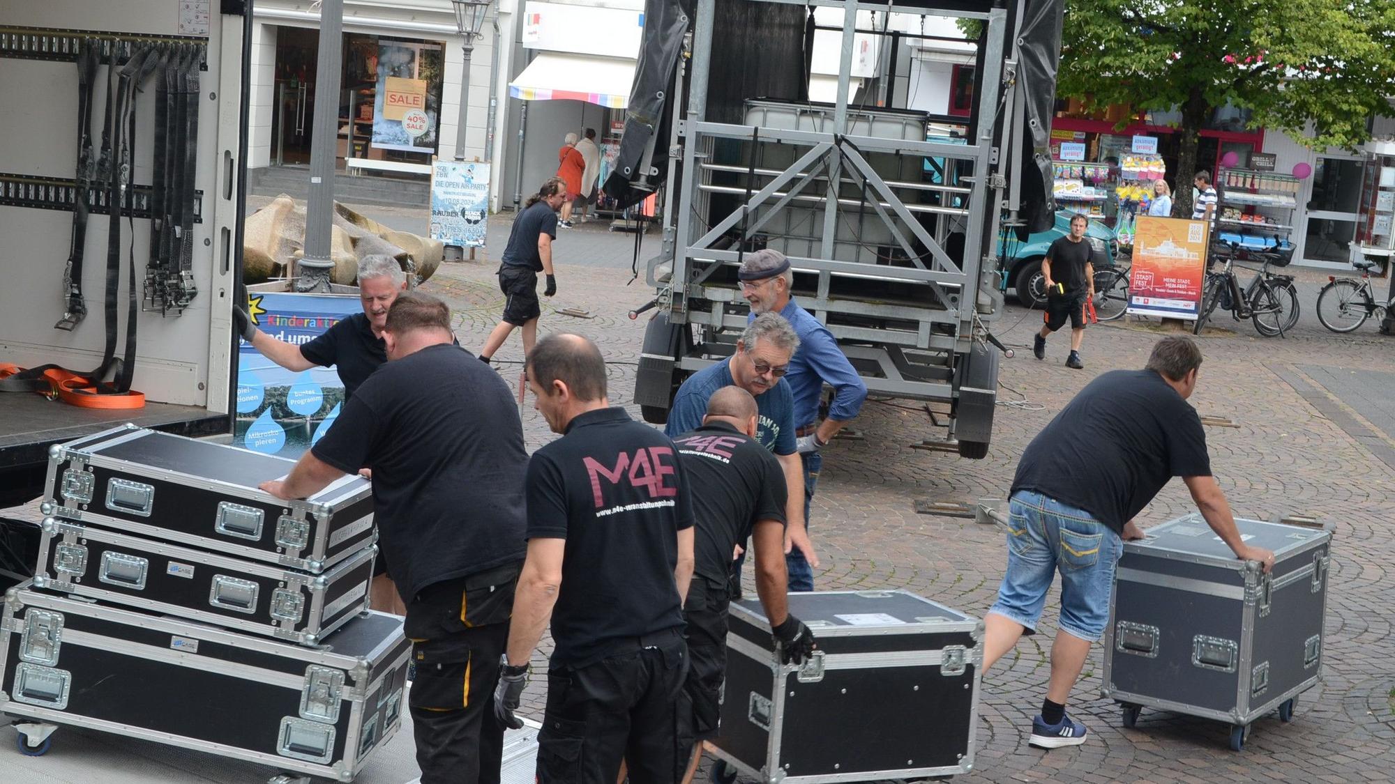 Männer entladen einen Lastwagen; sie rollen schwarze Boxen auf einen gepflasterten Platz.