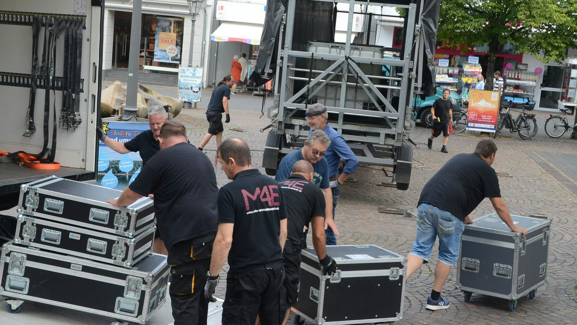Männer entladen einen Lastwagen; sie rollen schwarze Boxen auf einen gepflasterten Platz.
