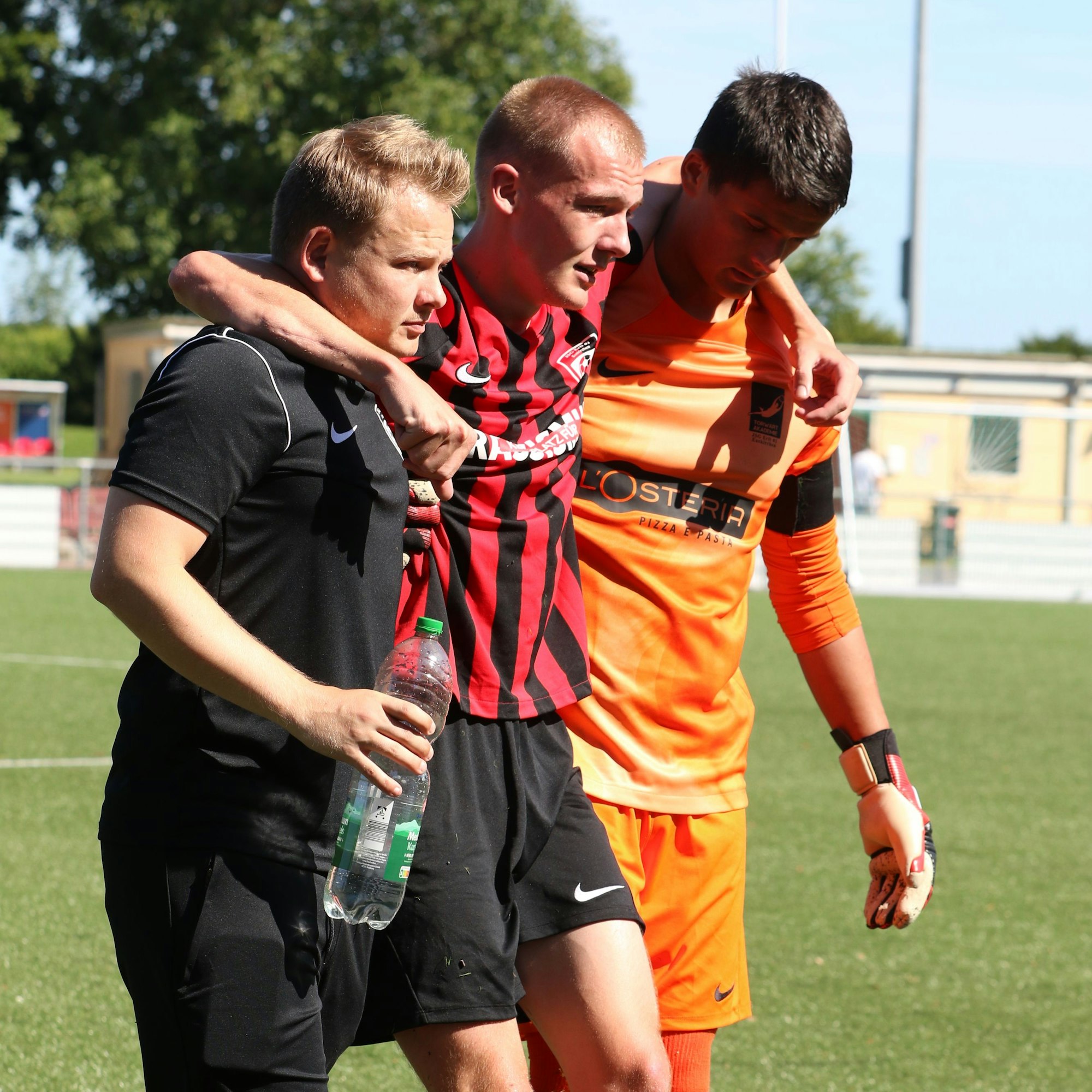 Ein verletzter Spieler wird von seinem Trainer und dem Torwart der Mannschaft gestützt.