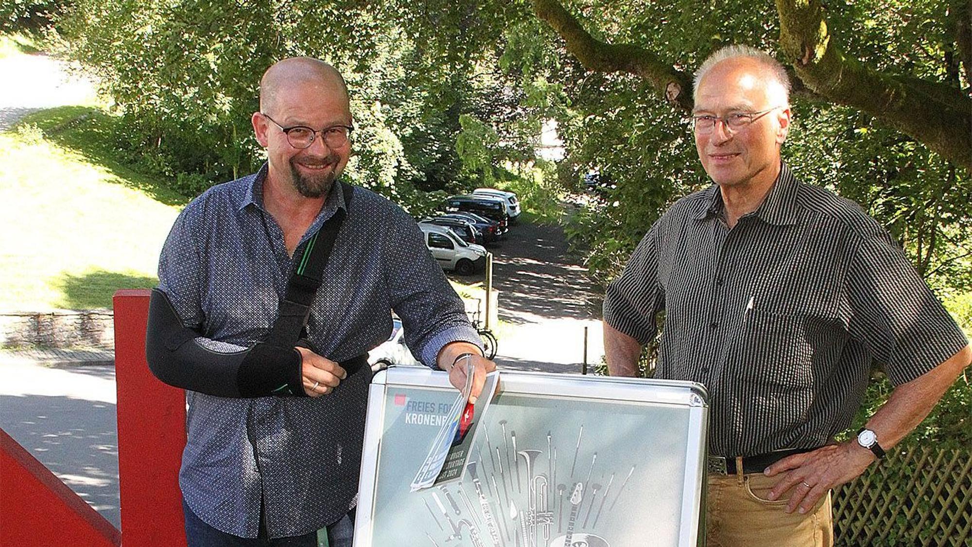 Zwei Männer, Martin Schöddert (links mit einer schwarzen Schulterschlinge) und Pit Igelmund, stehen an einem Plakataufsteller der Kronenburger Kunst- und Kulturtage.