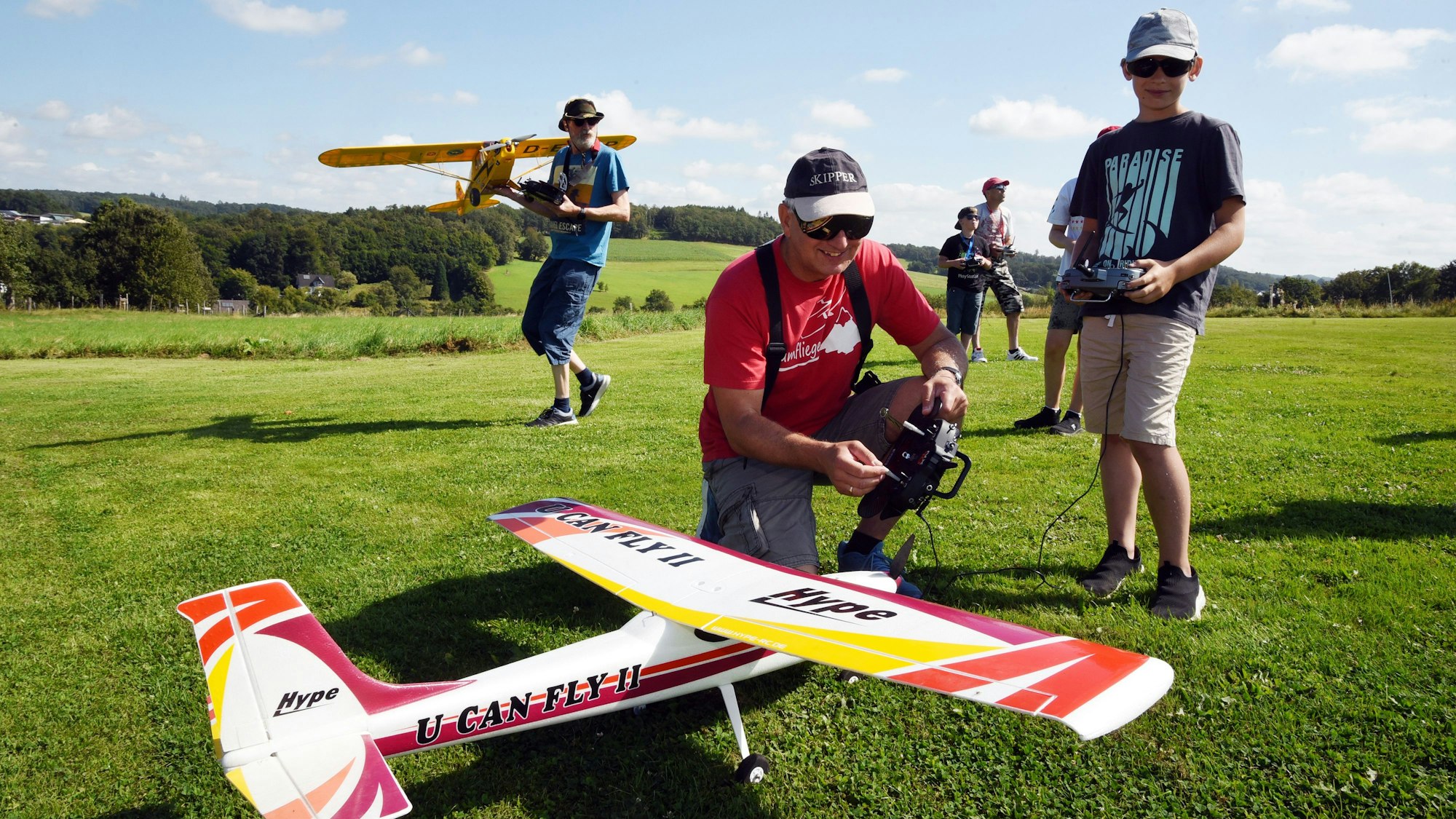 Ein Mann hockt neben einem Modellflugzeug. Er hält eine Fernbedienung in der Hand. Neben ihm steht ein Junge.