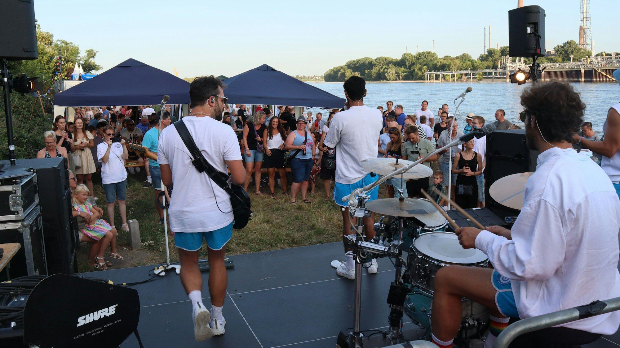 Eine Band in kurzen Hosen steht auf einer Bühne vor Publikum, dahinter fließt der Rhein.