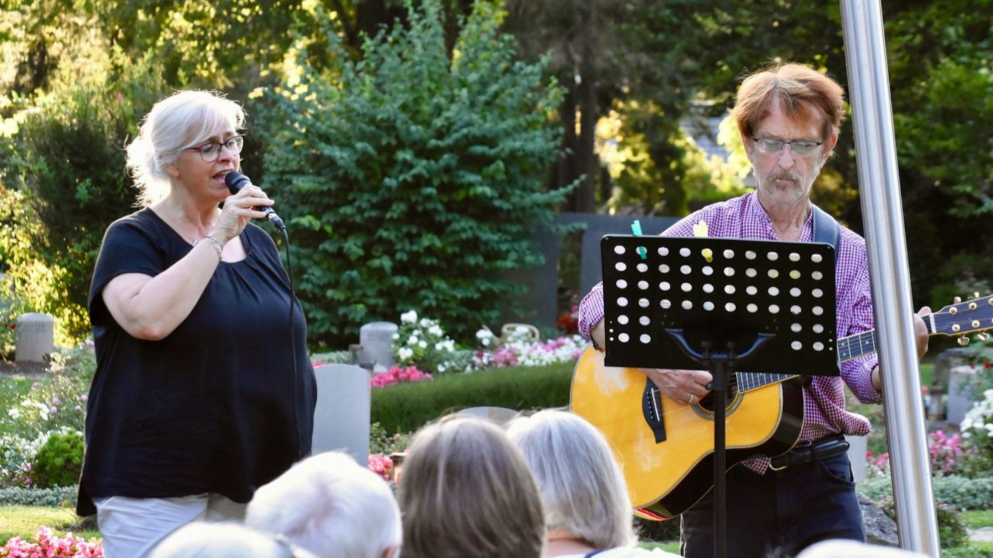Zwischen Gräbern singt eine Frau in ein Mikrfofon, neben ihr steht ein Mann und spielt Gitarre.