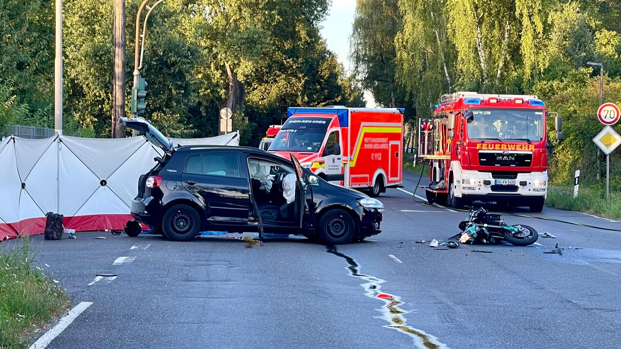 Ein Auto steht auf einer Straße vor mehreren Einsatzfahrzeugen der Feuerwehr, ein zerstörtes Motorrad liegt auf der Straße. Ein Sichtschutz ist hinter dem Pkw aufgebaut.