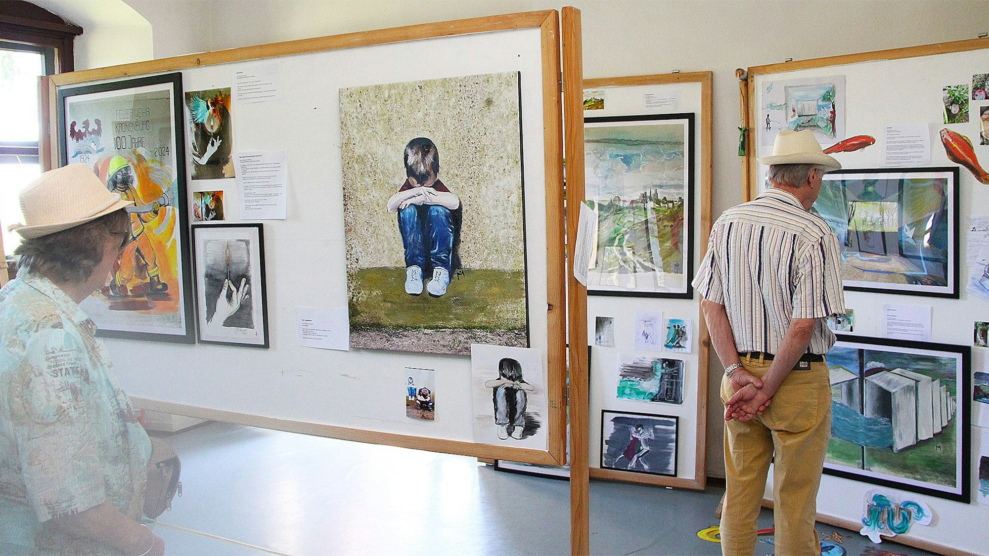 Eine Frau und ein Mann tragen Sommerhüte. Sie sind von hinten zu sehen, wie sie in einem Haus in Dahlem-Kronenburg Kunstwerke betrachten.