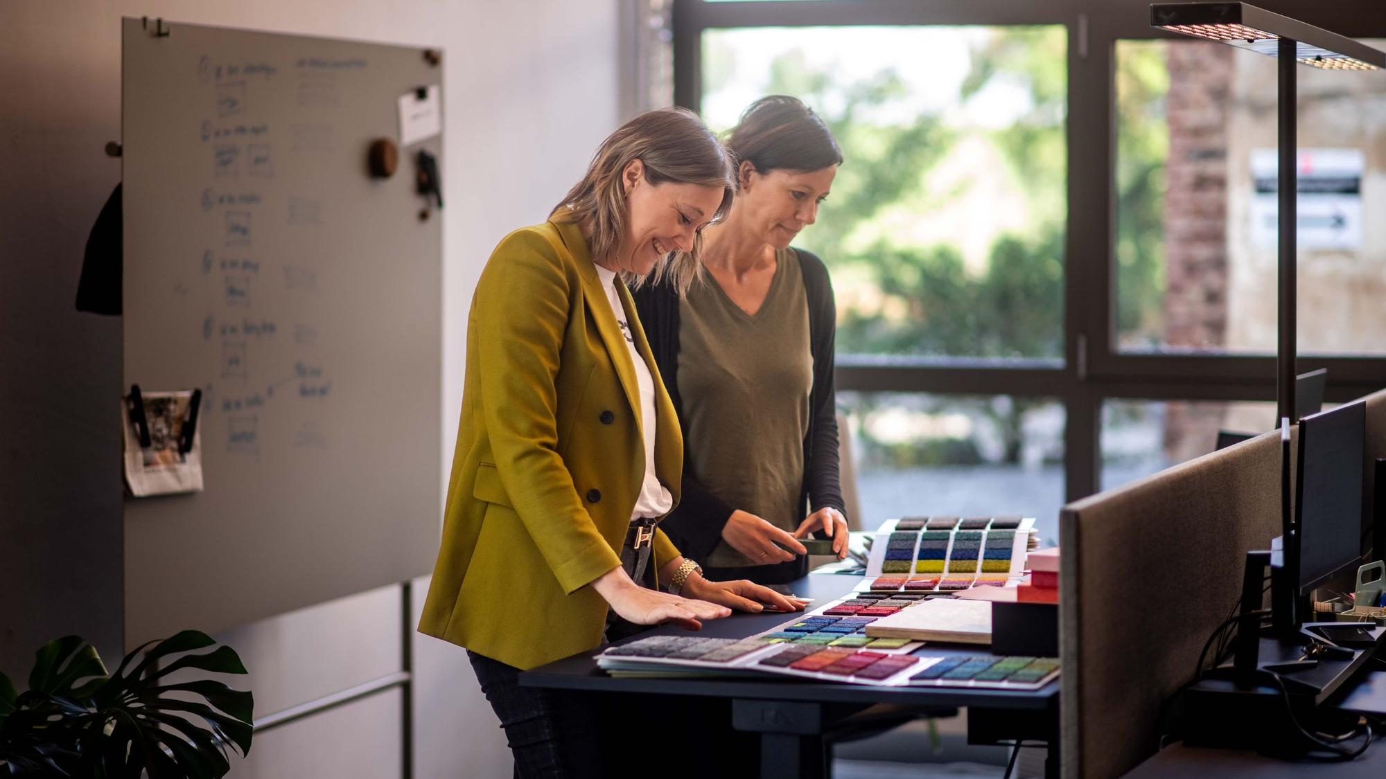 Nadia Stanke (l.) steht mit einer Mitarbeiterin am einem Tisch, auf dem Muster verschiedener Farben liegen.