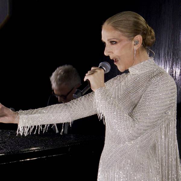Céline Dion hält einen Arm ausgestreckt während sie vor einem Pianisten auf der ersten Etage des Eiffelturms singt. Sie trägt ein weißes Perlenkleid von Dior.