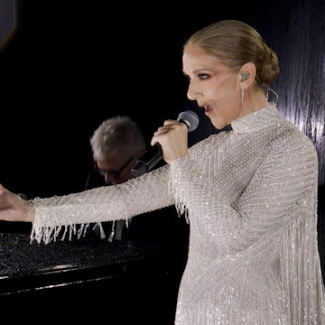 Céline Dion hält einen Arm ausgestreckt während sie vor einem Pianisten auf der ersten Etage des Eiffelturms singt. Sie trägt ein weißes Perlenkleid von Dior.