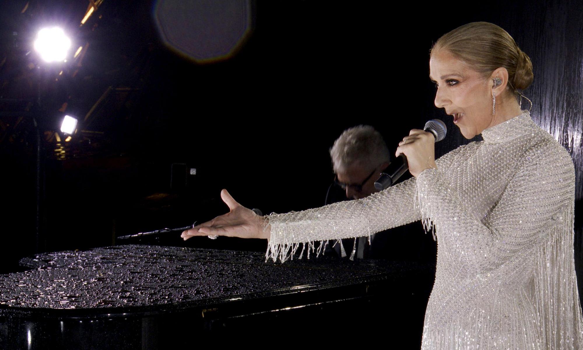 Céline Dion hält einen Arm ausgestreckt während sie vor einem Pianisten auf der ersten Etage des Eiffelturms singt. Sie trägt ein weißes Perlenkleid von Dior.