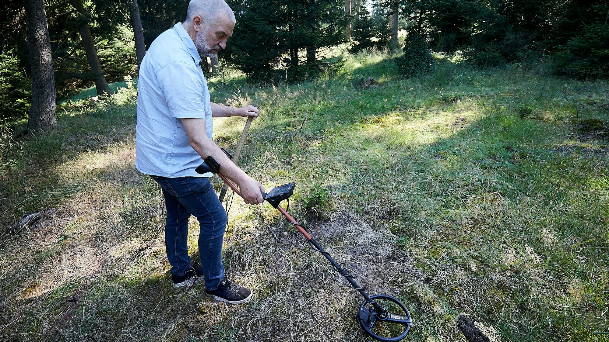 Frank Güth hält ein Metallsuchgerät in der Hand. Die Sonde hält er auf einer Wiese in einem Wald knapp über dem Boden.