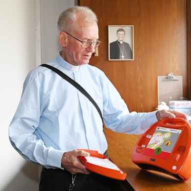 Pfarrer Franz Meurer zeigt einen Defibrillator in der Sakristei der Kirche Sankt Elisabeth in Köln. Im Hintergrund das Porträt des Kölner Erzbischofs, Kardinal Rainer Woelki