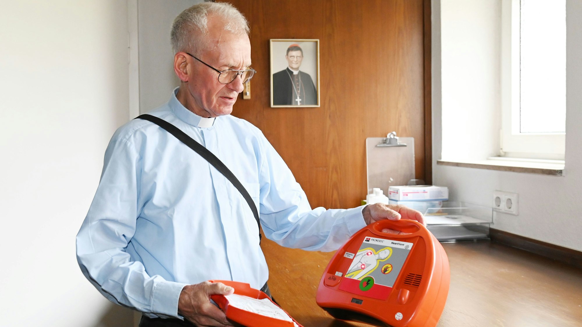 Pfarrer Franz Meurer zeigt einen Defibrillator in der Sakristei der Kirche Sankt Elisabeth in Köln. Im Hintergrund das Porträt des Kölner Erzbischofs, Kardinal Rainer Woelki