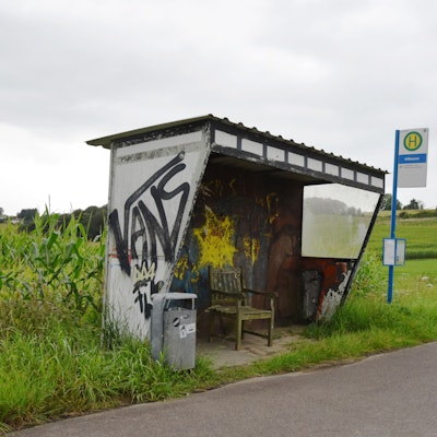 Eine mit Graffiti versehene Bushaltestelle steht mitten im Nirgendwo, drumherum nur Felder und weite Landschaften. Ein Haltestellenschild steht aus Sicht des Betrachtenden rechts von der Haltestelle. Links von der Haltestelle steht ein Mülleimer.