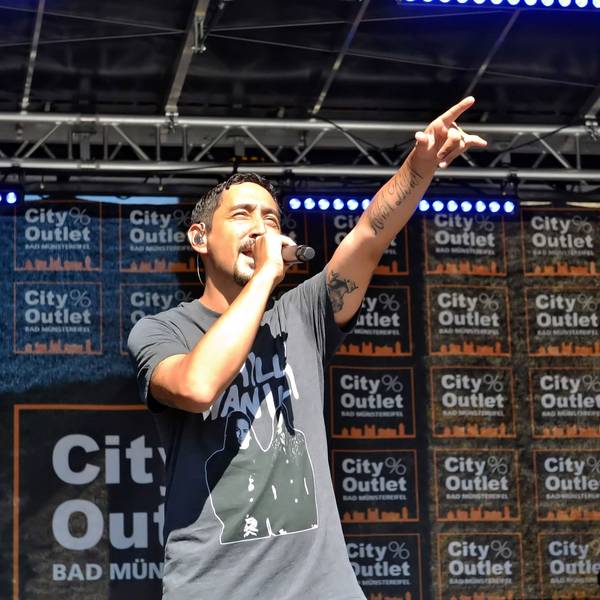 Eko Fresh wird auf der Bühne von der Sonne geblendet und streckt den linken Zeigefinger in die Höhe. Im Hintergrund steht auf vielen Bannern City Outlet Bad Münstereifel.