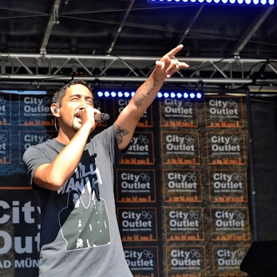 Eko Fresh wird auf der Bühne von der Sonne geblendet und streckt den linken Zeigefinger in die Höhe. Im Hintergrund steht auf vielen Bannern City Outlet Bad Münstereifel.