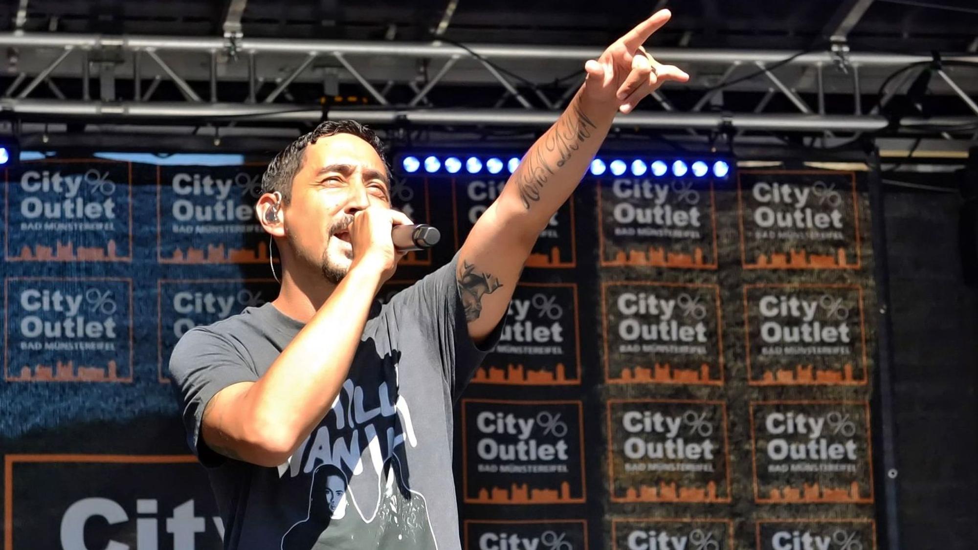 Eko Fresh wird auf der Bühne von der Sonne geblendet und streckt den linken Zeigefinger in die Höhe. Im Hintergrund steht auf vielen Bannern City Outlet Bad Münstereifel.