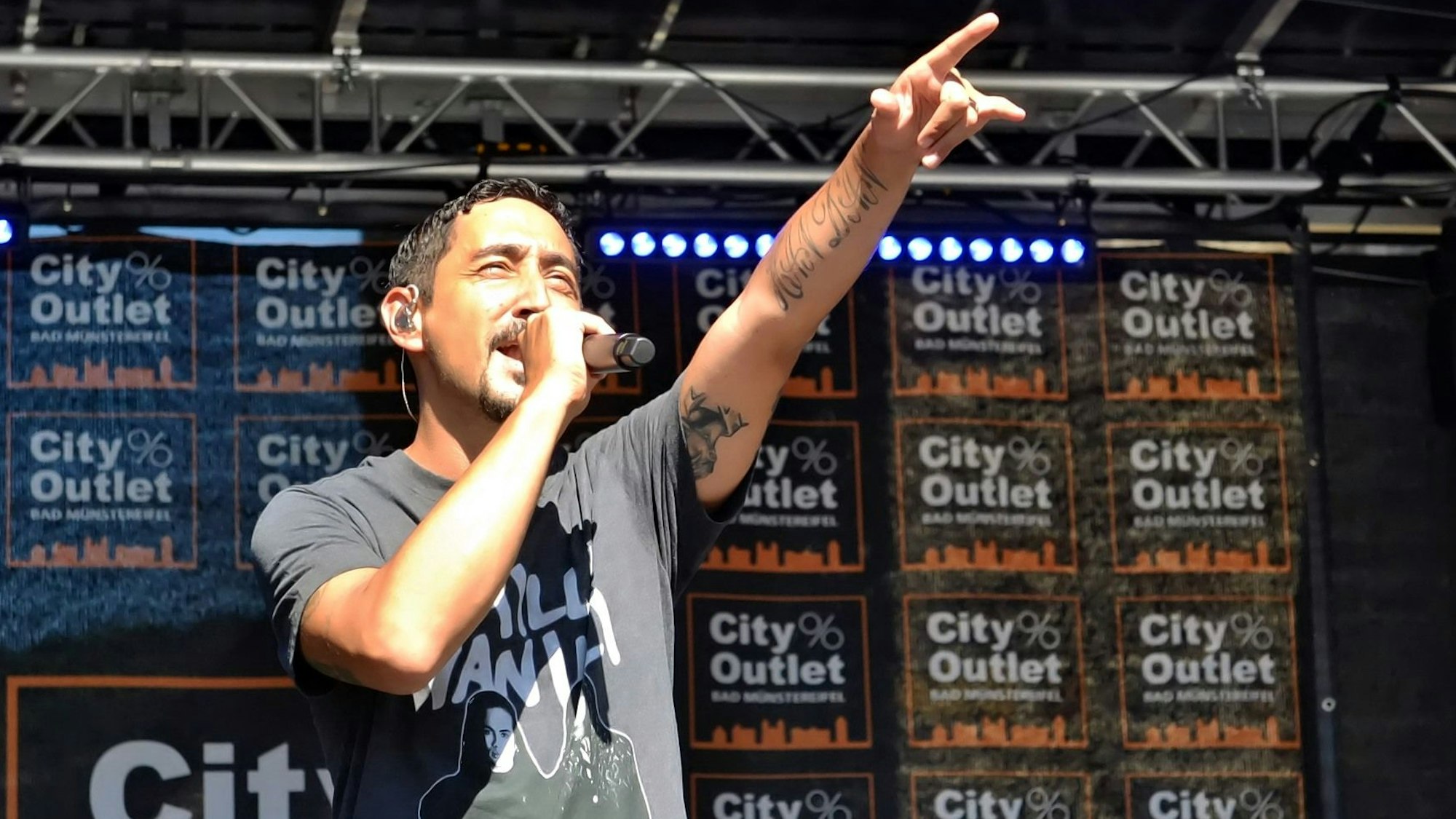 Eko Fresh wird auf der Bühne von der Sonne geblendet und streckt den linken Zeigefinger in die Höhe. Im Hintergrund steht auf vielen Bannern City Outlet Bad Münstereifel.