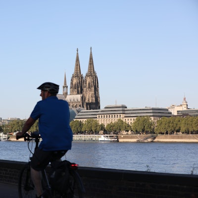 Radfahrer fahren am Rheinufer entlang, im Hintergrund ist der Kölner Dom zu sehen