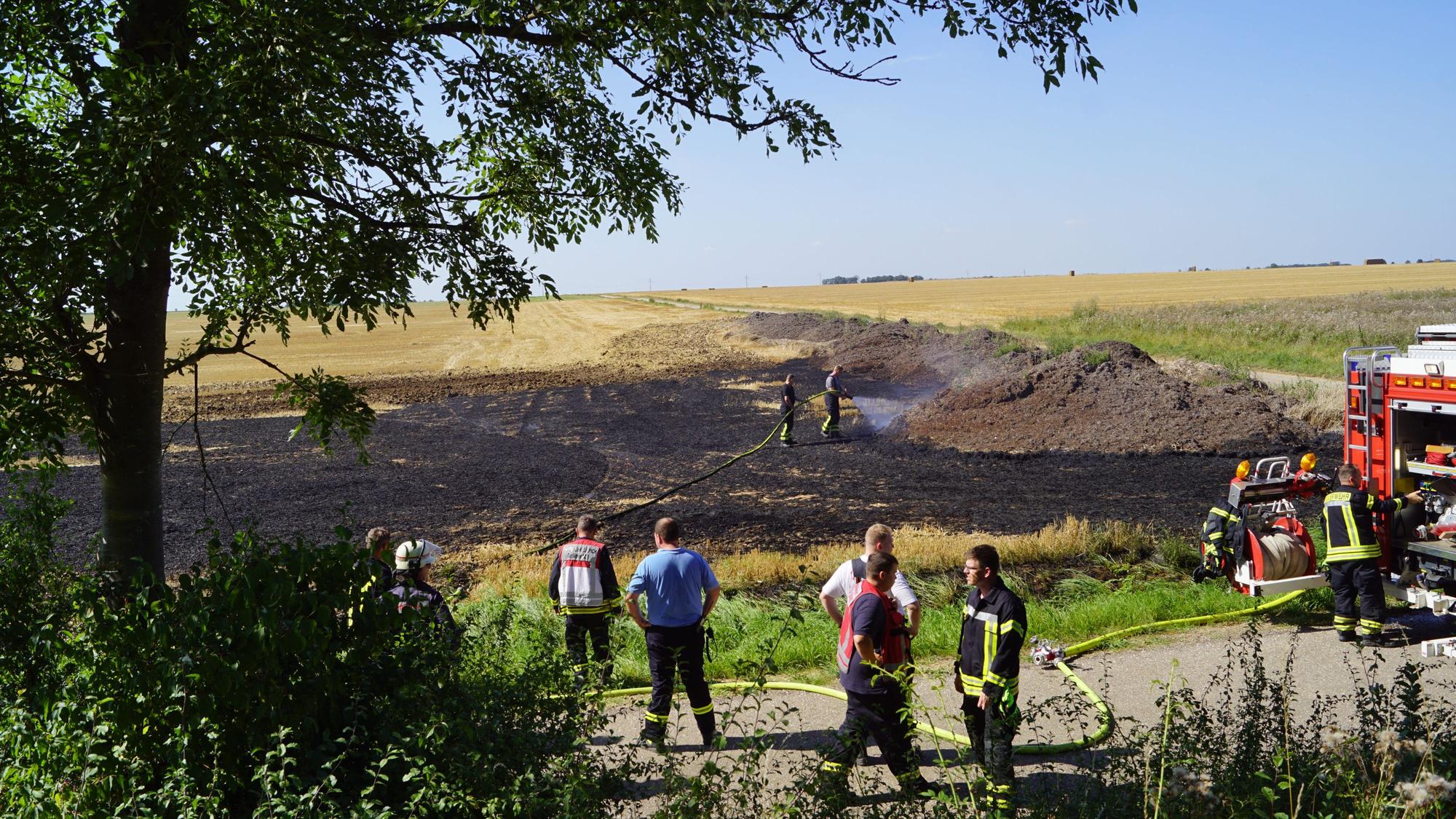Auf einem Feld qualmen Strohballen. Rechts im Bild ein Feuerwehrauto, Feuerwehrleute löschen mit einem Schlauch.