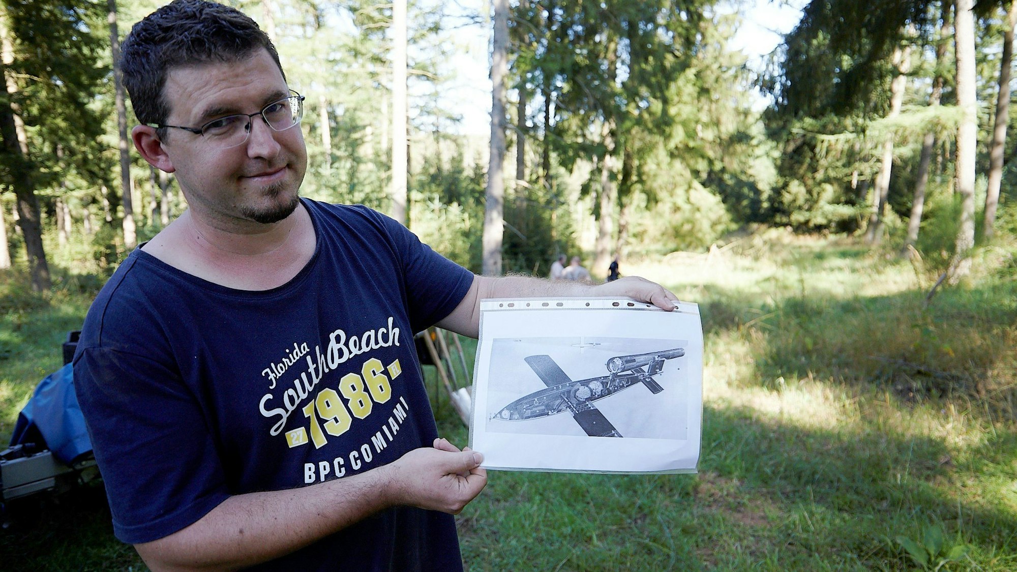 Daniel Rechziger steht in einem Wald und hält ein Foto einer V1-Flugbombe aus dem Zweiten Weltkrieg in der Hand.