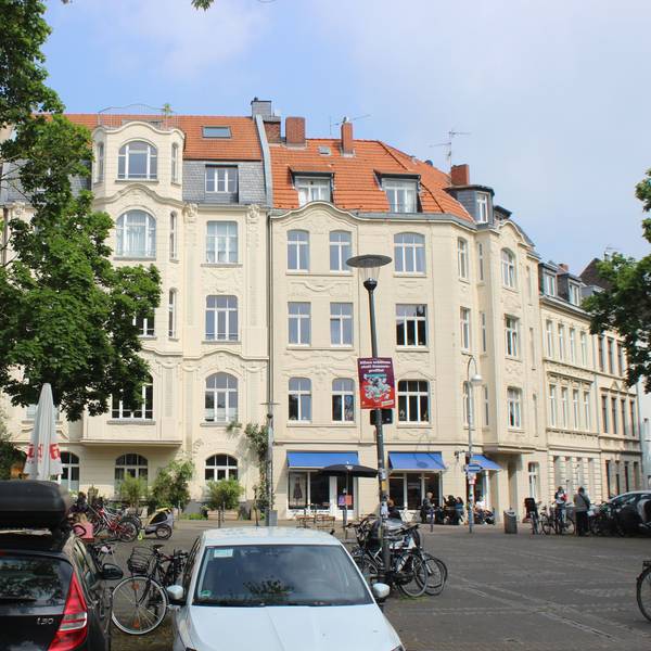 Zu sehen sind drei Jugendstil-Gebäude. Davor sitzen Menschen auf dem Schillplatz in Nippes.
