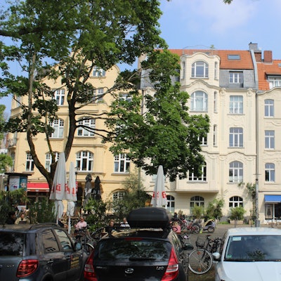 Zu sehen sind drei Jugendstil-Gebäude. Davor sitzen Menschen auf dem Schillplatz in Nippes.