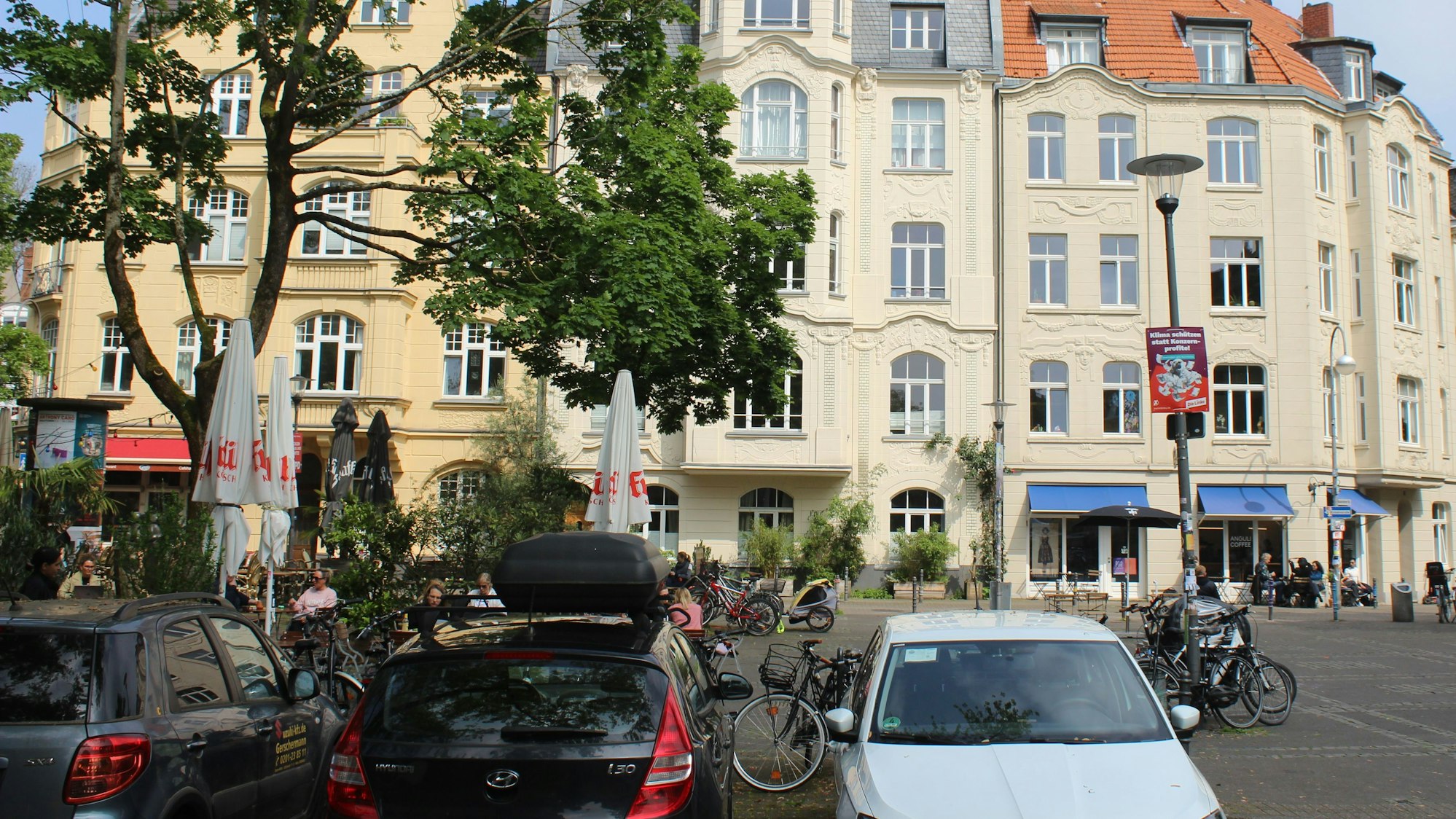 Zu sehen sind drei Jugendstil-Gebäude. Davor sitzen Menschen auf dem Schillplatz in Nippes.