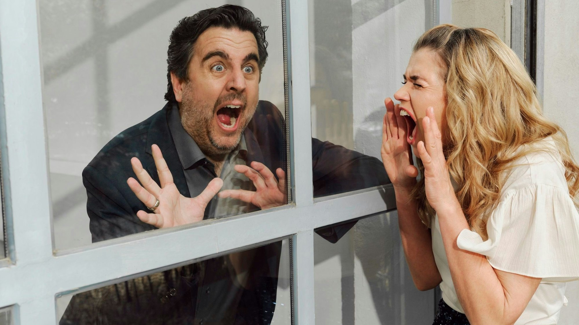 Anke Engelke und Bastian Pastewka befinden sich auf je einer Seite eines Fensters und mimen Panik.