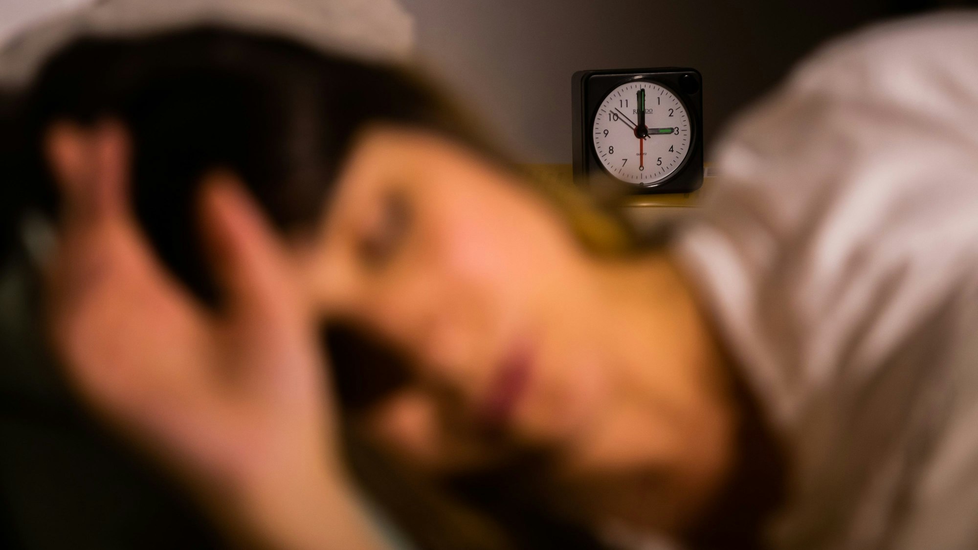 Eine Frau schläft neben einem Wecker, der auf drei Uhr zeigt (gestellte Szene).