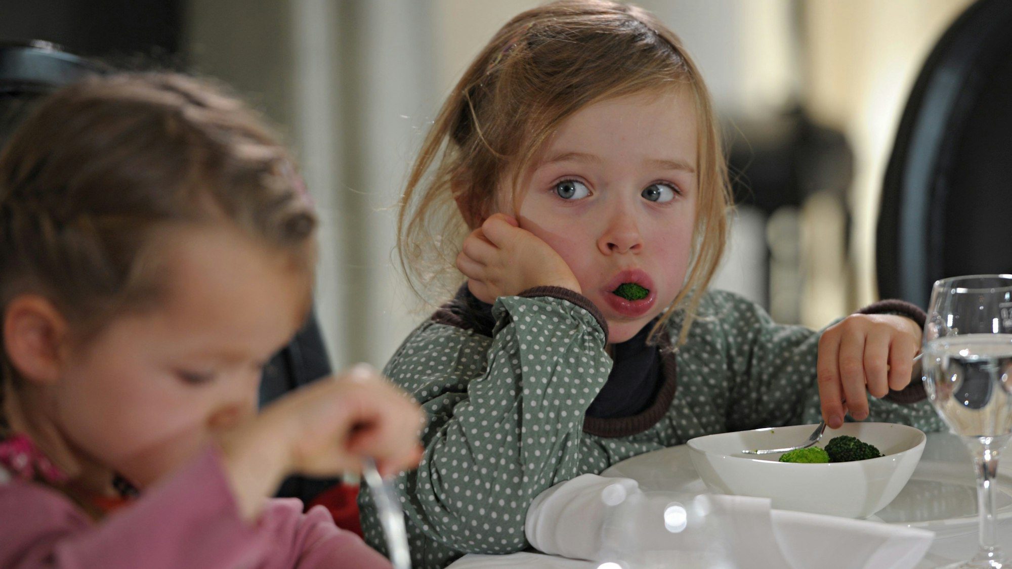 Zwei kleine Mädchen sitzen an einem mit Tischtuch, Stoffservietten, weißem Geschirr und Stilgläsern gedeckten Tisch. Eines der Mädchen hat eine Gabel in der Hand, das andere stützt ihren Kopf mit einer Hand ab, während die andere Hand ein Stück Brokkoli aus einer Schüssel nimmt. In ihrem geöffneten Mund ist ein weiteres großes Stück Brokkoli sichtbar.