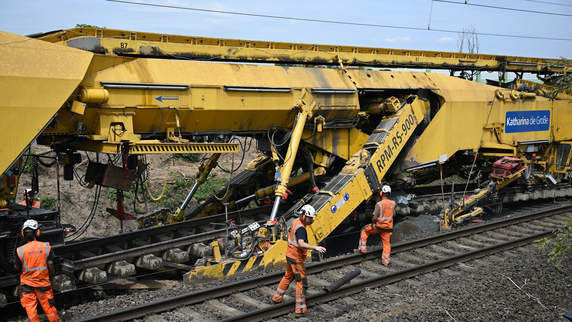 Die fast 200 Meter lange Spezialmaschine RPM-RS-900, genannt ·Katharina die Große·, ist bei der Generalsanierung der Riedbahn im Einsatz. Ein großes Gerät auf Schienen, davor sind Arbeiter in Montur zu sehen.