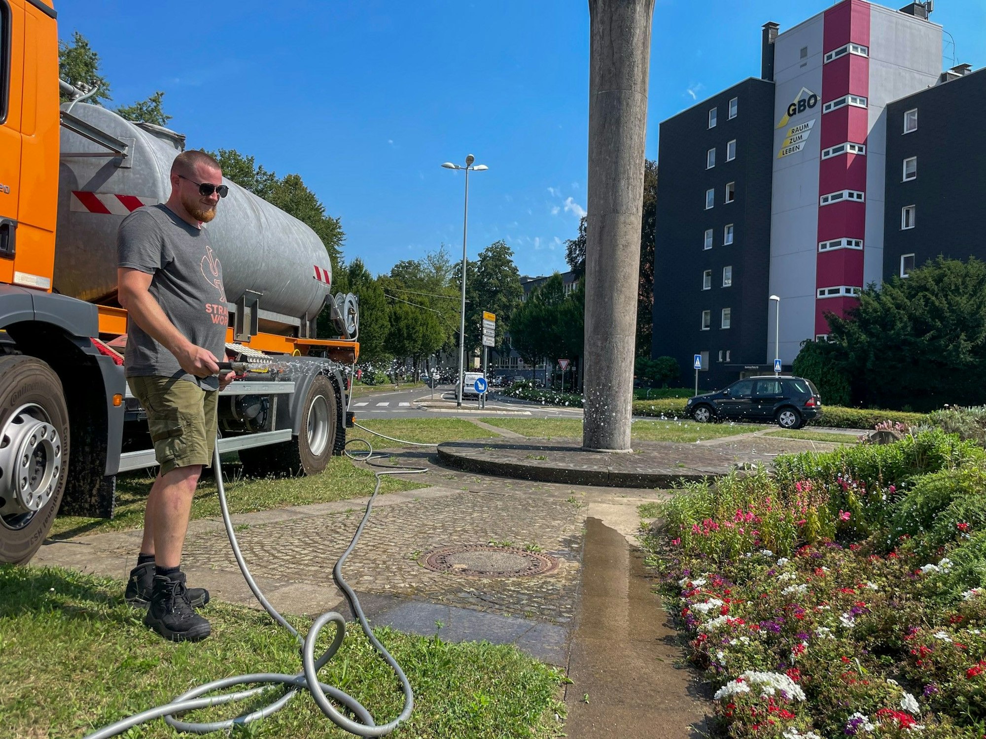 Ein Mann wässert ein städtisches Beet, dahinter ist Wohnbebauung zu sehen.